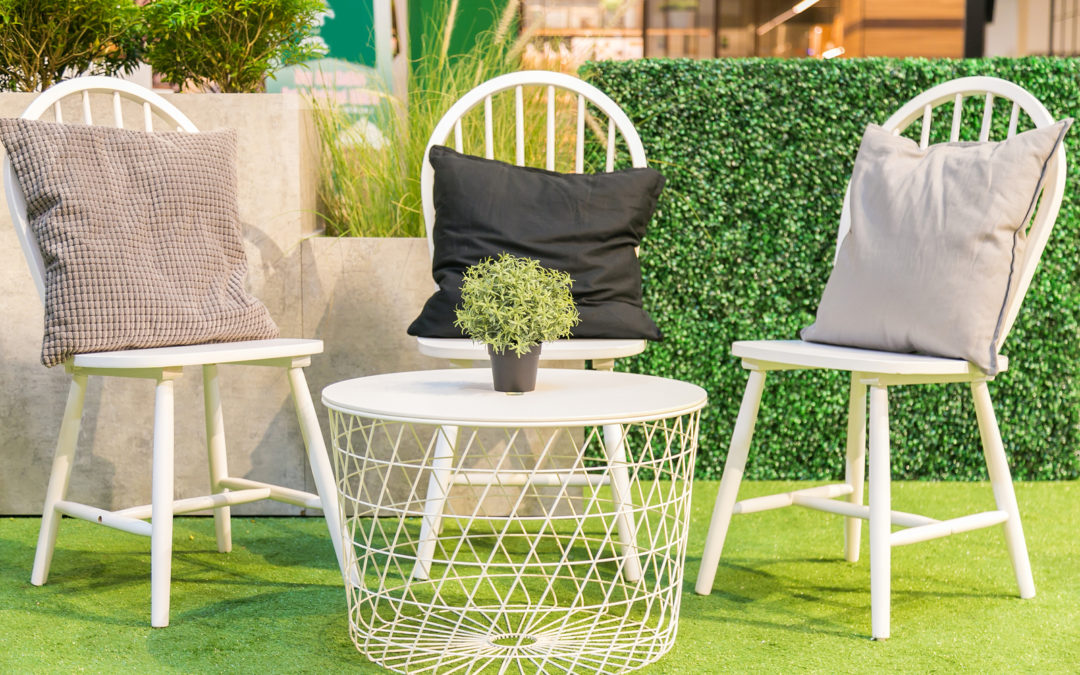 A Pillows on a white wooden chairs and a Artificial tree in pot on a white table on artificial grass.